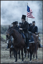 Men in period clothing riding horses through smoke, one man wearing a top hat and holding an American flag.