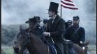 A man in a black top hat and suit rides a horse past soldiers under a cloudy sky with an American flag.