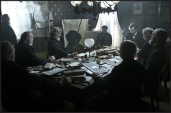 A group of men in dark clothing sit around a large table covered in books and papers in a dimly lit room.