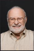 A smiling older man with glasses and a white beard wearing a striped button-down shirt.