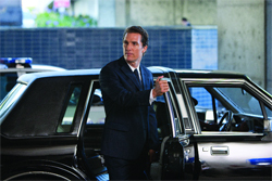 A man in a dark suit steps out of a black sedan near a police vehicle.