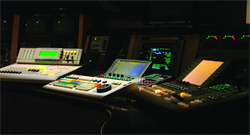 Multiple illuminated control panels and monitors in a dark broadcasting or production studio.