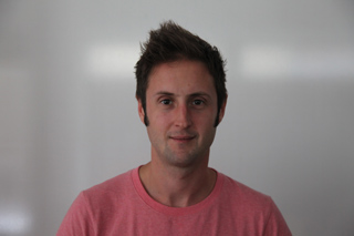 A man with short brown hair and sideburns wearing a pink t-shirt against a plain white background.