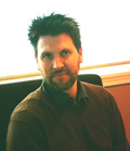 A man with dark hair and a beard wearing a brown sweater sits in front of a bright window.