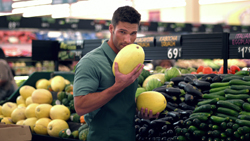 A man in a green shirt holds and smells a large yellow melon in a produce aisle.