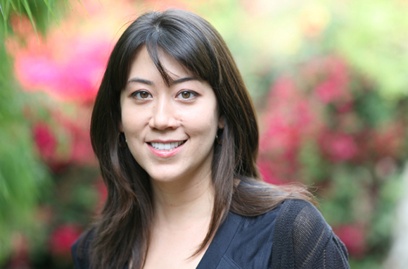 A smiling woman with long dark hair posing outdoors against a soft, colorful floral background.
