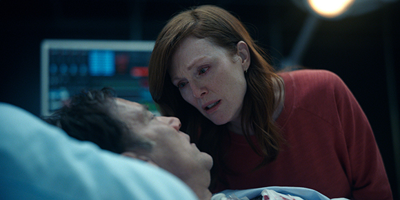 A woman with red hair leans over a man lying in a hospital bed in a dimly lit room.