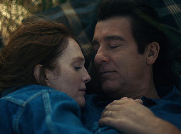 A man and a woman with red hair lie close together in a dimly lit setting with their eyes closed.