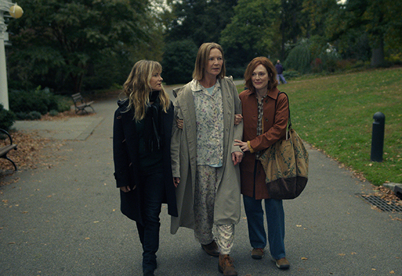 Three women walk side-by-side along a paved park path lined with trees and grass.