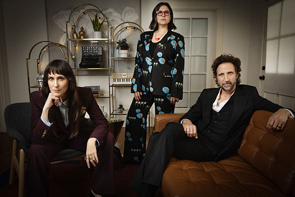 Three people in fashionable attire pose in a room with a gold shelving unit and a leather sofa.