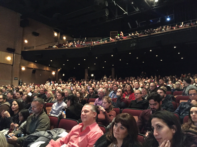 A full theater audience is seated in rows, with some people in the balcony above. The lighting is dim.