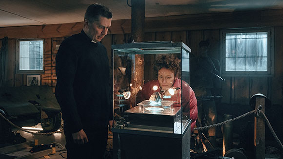 A man in a priest's collar stands beside a woman looking into a glowing glass display case in a dim room.