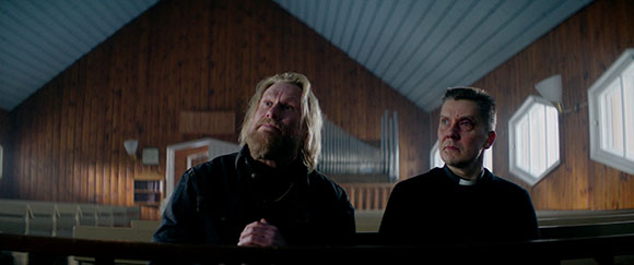 A man with long blonde hair and a beard sits next to a man in a clerical collar inside a wooden building.