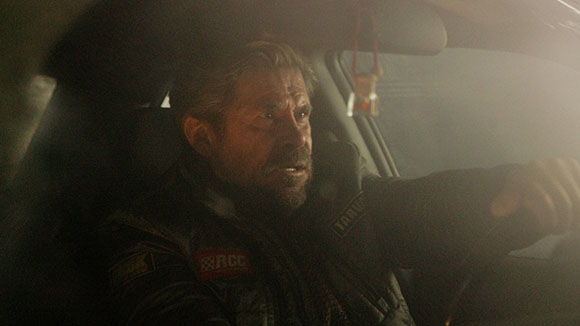 A bearded man with a focused expression sits in the driver's seat of a car in dim, hazy lighting.
