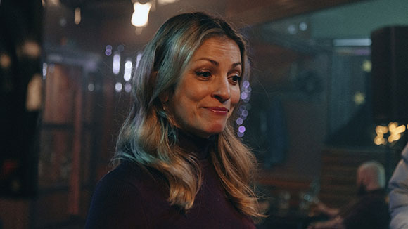 A blonde woman smiles warmly in a dimly lit indoor setting.