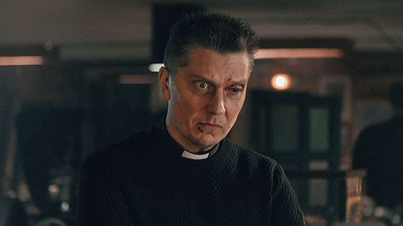 A man wearing a black sweater and a white clerical collar looks to the side with a worried expression.