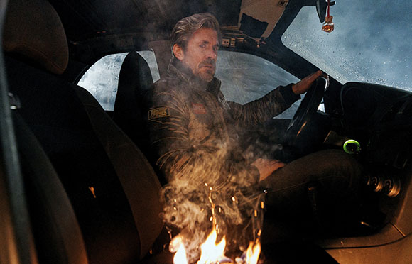 A man with light hair sits in a dark vehicle next to a small fire with rising smoke.