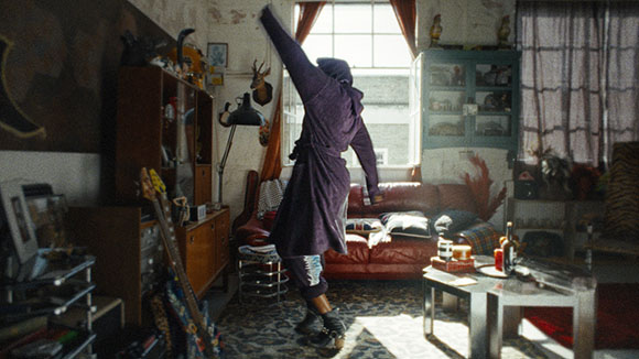 A person in a long purple robe dances in a cluttered, sunlit living room near a red sofa.
