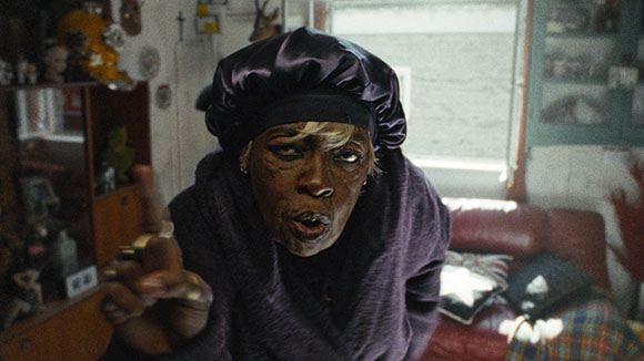 A person in a purple bonnet and dark clothing leans forward, pointing toward the camera in a dimly lit room.