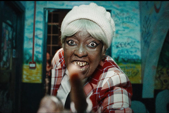 A woman with white hair and a white beanie stares intensely with a wide-eyed, grimacing expression.