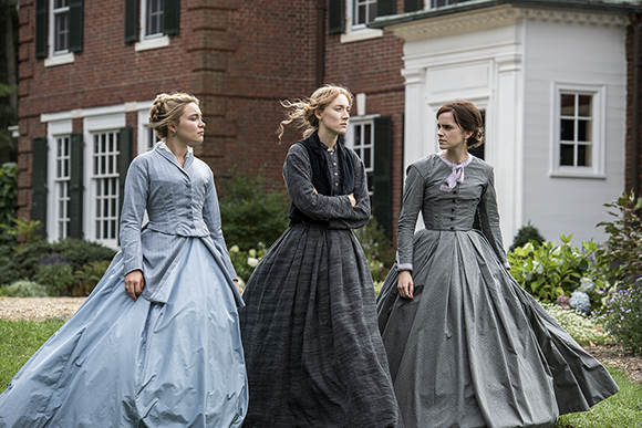 Three women in long, period-style dresses stand in a garden before a large brick and white house.