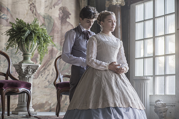 A young man in a waistcoat helps a young woman in a white period gown stand by a large window.