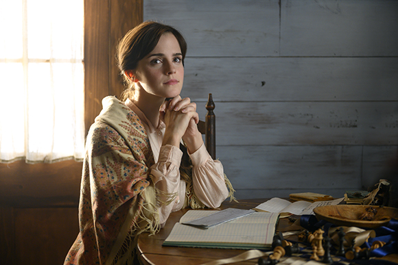 A woman in period clothing sits at a table with books and writing materials near a window.