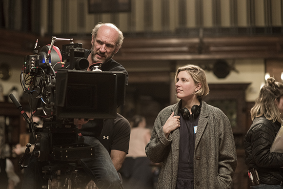A man operates a large professional movie camera while a woman in a grey coat watches nearby.