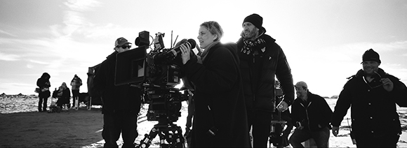 A film crew operates a large camera on a tripod against a bright sky.