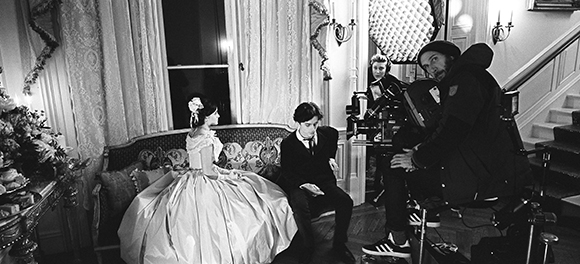 Black and white photo of a film set featuring actors in period clothing and a camera operator.