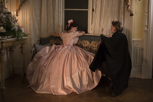 A woman in a voluminous pink ballgown sits on a patterned sofa as a stylist works beside her.