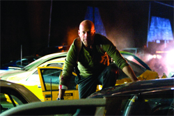 A bald man in a green long-sleeve shirt crouches between yellow and dark cars at night.