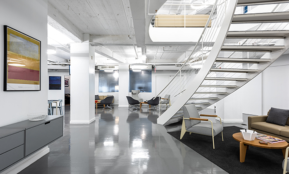 A white spiral staircase descends into a bright, modern open-plan office with grey floors and lounge seating.