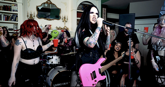 A woman with long black hair sings into a microphone while holding a pink electric guitar in a crowded room.