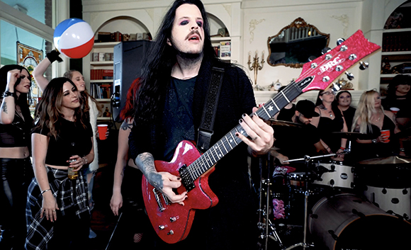 A man with long black hair plays a red electric guitar while people stand in the background.
