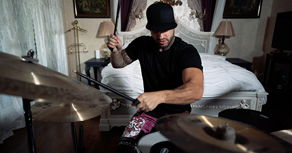 A man in a black t-shirt playing a drum kit in a bedroom with a white bed in the background.