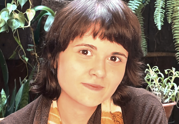 A young woman with dark, wavy hair and bangs looks toward the camera against a background of green foliage.