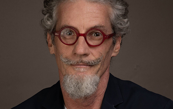 A man with curly grey hair, red round glasses, and a grey goatee looking at the camera.