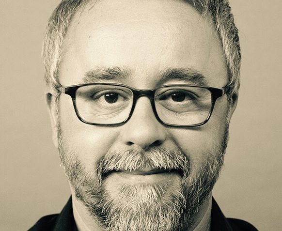 Close-up portrait of a man with short hair, glasses, and a salt-and-pepper beard looking at the camera.