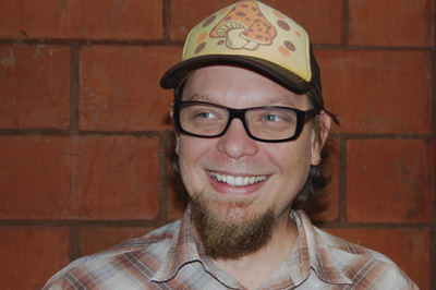 Smiling man with glasses, a goatee, and a yellow hat featuring pizza and mushroom illustrations.
