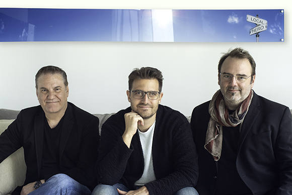 Three men sitting side-by-side on a light-colored couch against a white wall.