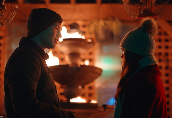 A man in a beanie and a woman in a pom-pom hat face each other by a glowing outdoor fire pit.