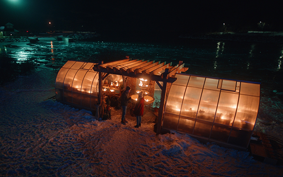 A warm, lit translucent structure sits on a snowy beach next to dark water at night.
