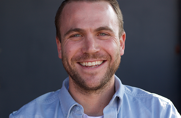 A smiling man with short brown hair and a beard wearing a light blue button-down shirt.