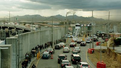 Vehicles and people queue in a line next to a tall concrete wall under a cloudy sky.