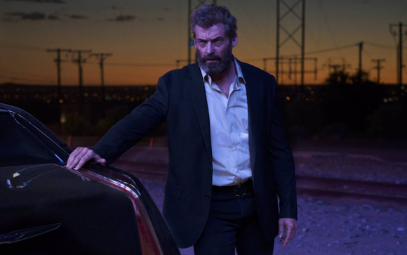 A bearded man in a dark suit and white shirt stands next to a car against a twilight sky with power lines.