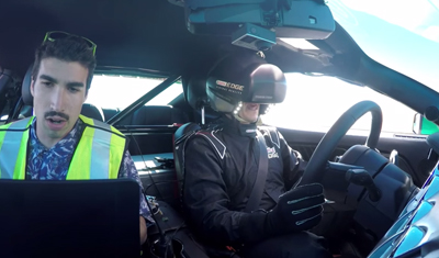 A man in a neon safety vest watches a laptop while a driver in a racing suit uses a VR headset in a car.