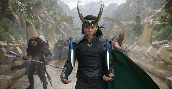 A man in black armor and a gold horned helmet stands amidst a battle in a rocky mountain pass.