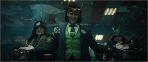 A man in a green vest and horned headpiece stands with arms outstretched in a dark setting.