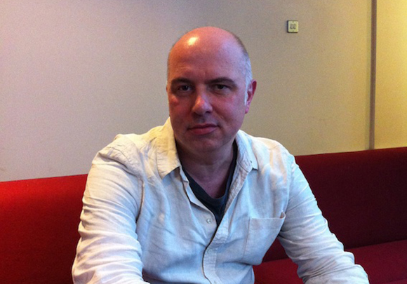 A man with a shaved head wearing a white button-down shirt sits on a red couch.